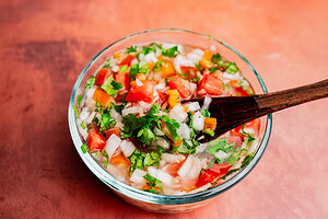 A glass bowl filled with freshly made pico de gallo, featuring chopped tomatoes, onions, cilantro, and peppers, is enhanced with a touch of aji picante. A wooden spoon rests inside.