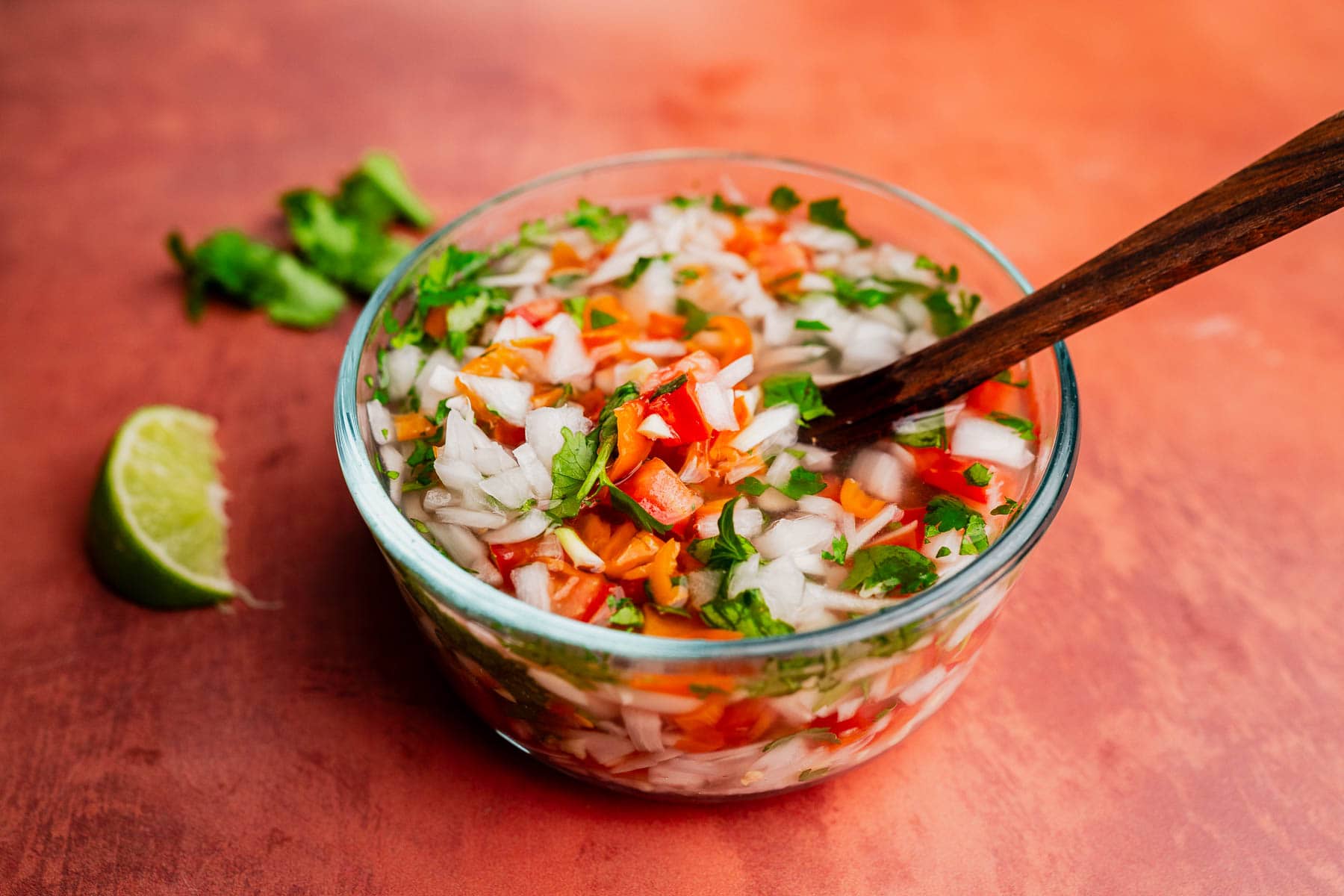A glass bowl of chopped tomatoes, onions, cilantro, and peppers with a wooden spoon sits next to a lime wedge on an orange surface, ready for a splash of zesty aji picante.
