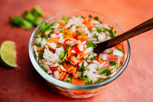 A glass bowl of chopped vegetables including onions, tomatoes, cilantro, and carrots with a wooden spoon on a reddish surface. A lime wedge and a hint of aji picante are in the background.
