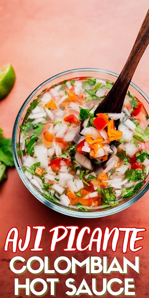 A bowl of aji picante, Colombian hot sauce with chopped onions, peppers, cilantro, and a wooden spoon sits on a reddish surface.
