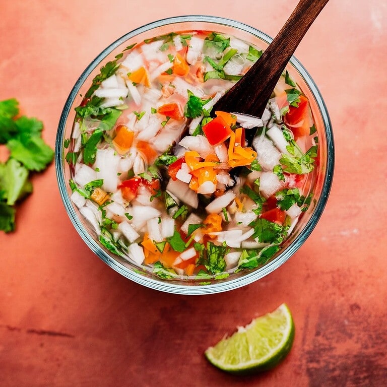 A glass bowl of chopped onions, tomatoes, cilantro, and peppers mixed with liquid for aji picante sits with a wooden spoon inside. A lime wedge and fresh cilantro rest on the reddish surface alongside.