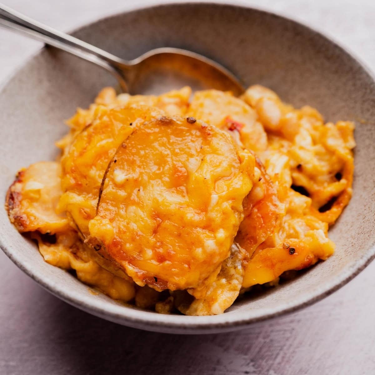 A bowl of cheesy buffalo potato casserole with a serving spoon, showing layers of sliced potatoes covered in melted cheese.