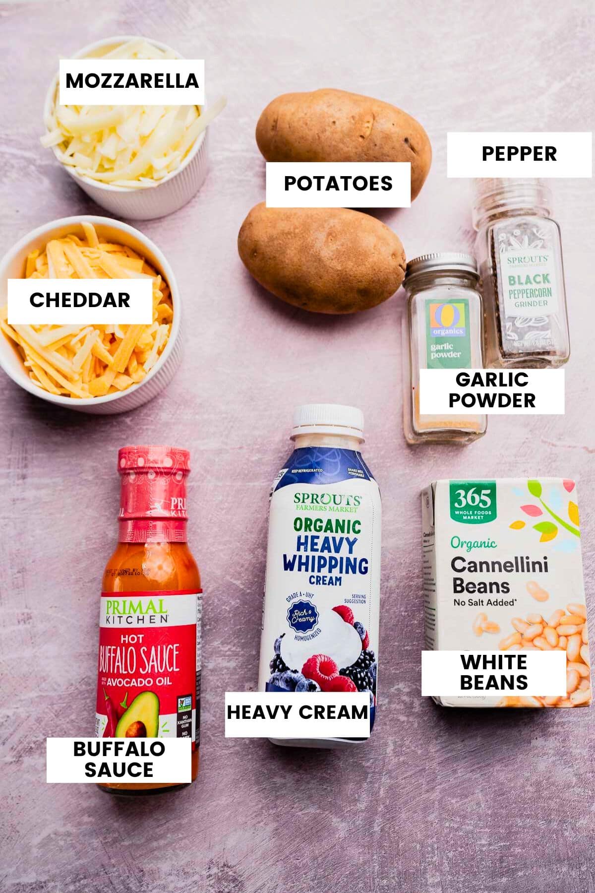 Various labeled ingredients are arranged on a purple surface for a buffalo potato casserole, including mozzarella, cheddar, potatoes, pepper, garlic powder, buffalo sauce, heavy cream, and white beans.