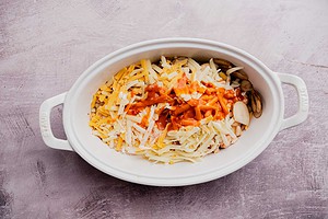 An oval white baking dish containing sliced mushrooms, shredded cheese, and a drizzle of orange sauce brings a bold twist to this buffalo potato casserole on a light surface.