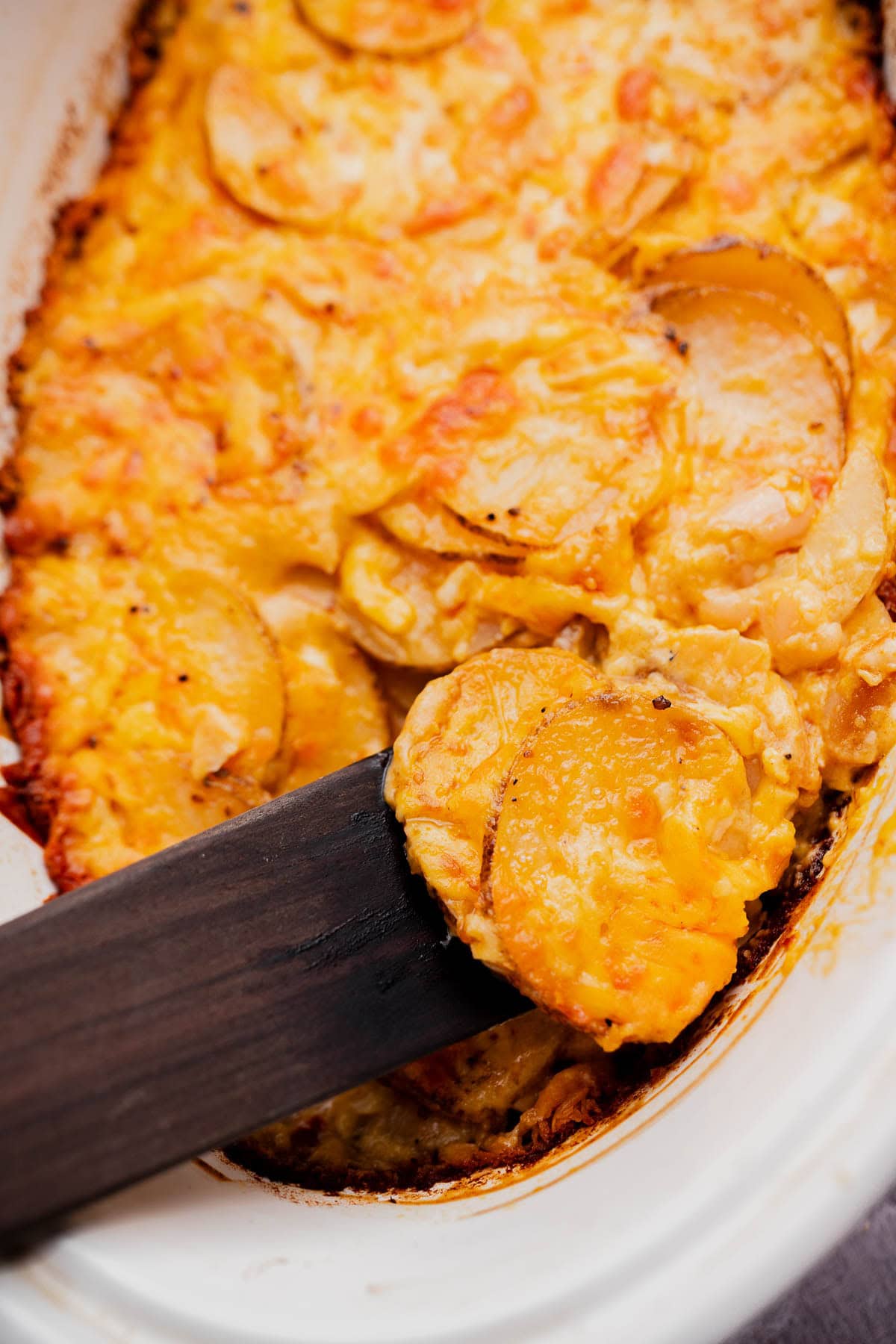 A serving of buffalo potato casserole is being lifted out of a white baking dish with a black spatula, showcasing its cheesy goodness.