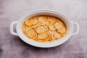 Oval white casserole dish filled with sliced potatoes and shredded cheese in a creamy buffalo potato casserole sauce, placed on a light textured surface.