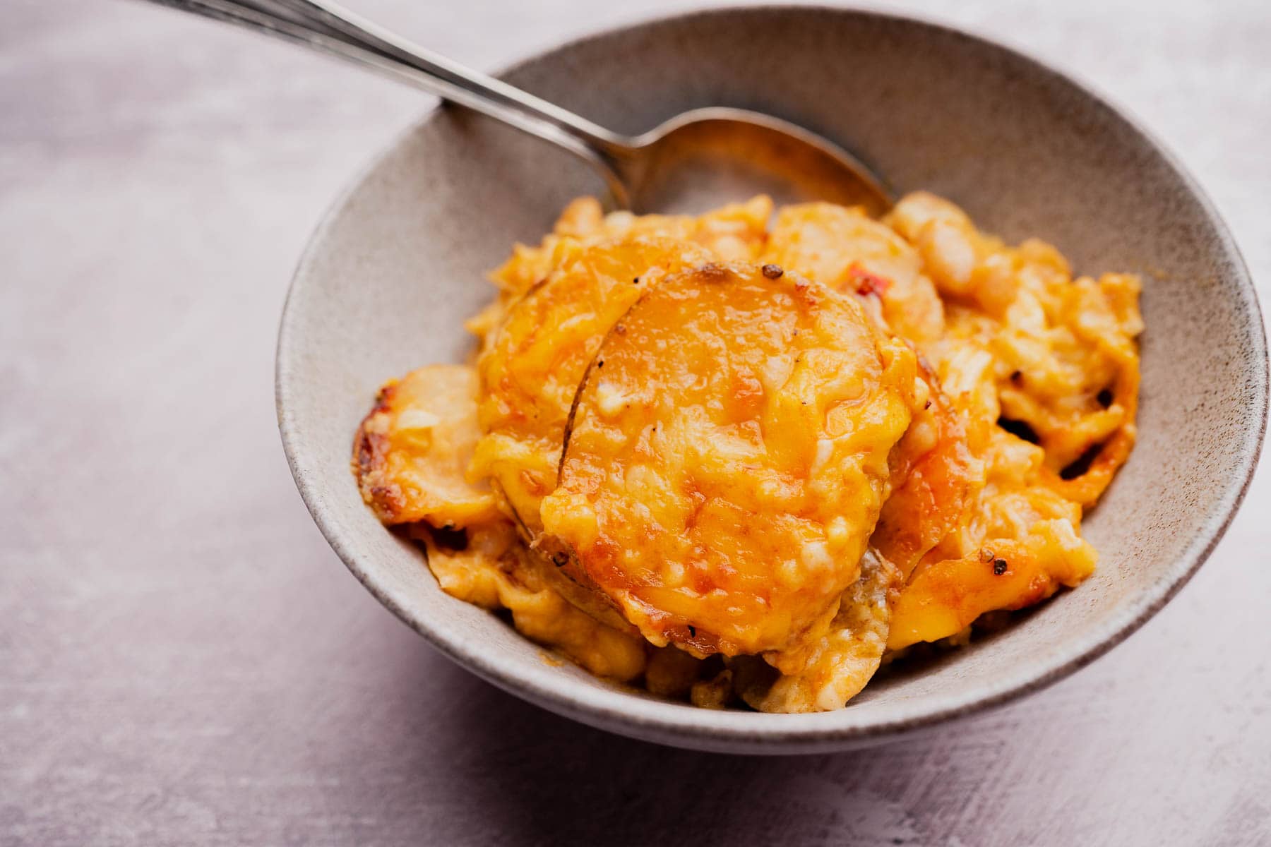 A bowl filled with cheesy buffalo potato casserole, topped with melted golden cheese, with a spoon resting in the bowl.