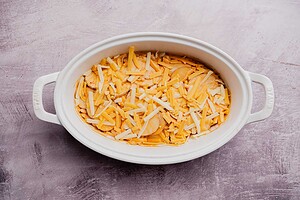 An oval white baking dish filled with sliced potatoes and shredded cheese, this buffalo potato casserole sits on a light textured surface.