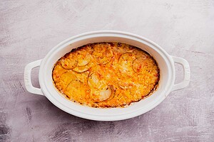 Oval white baking dish containing a golden-brown buffalo potato casserole, with visible layers of sliced potatoes and melted cheese on top.