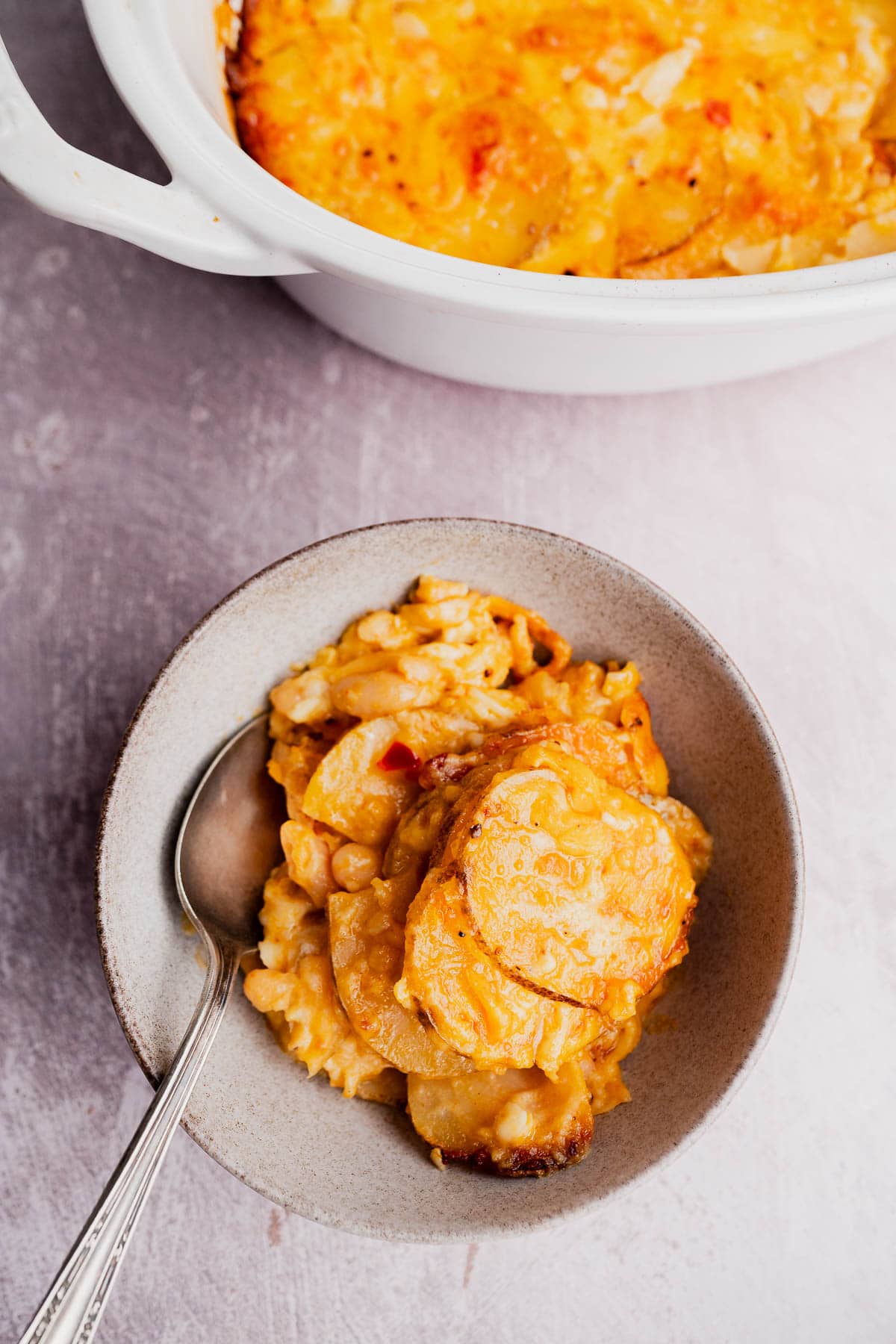 A bowl of cheesy scalloped potatoes with a spoon, next to a white baking dish filled with more buffalo potato casserole, sits on a light surface.