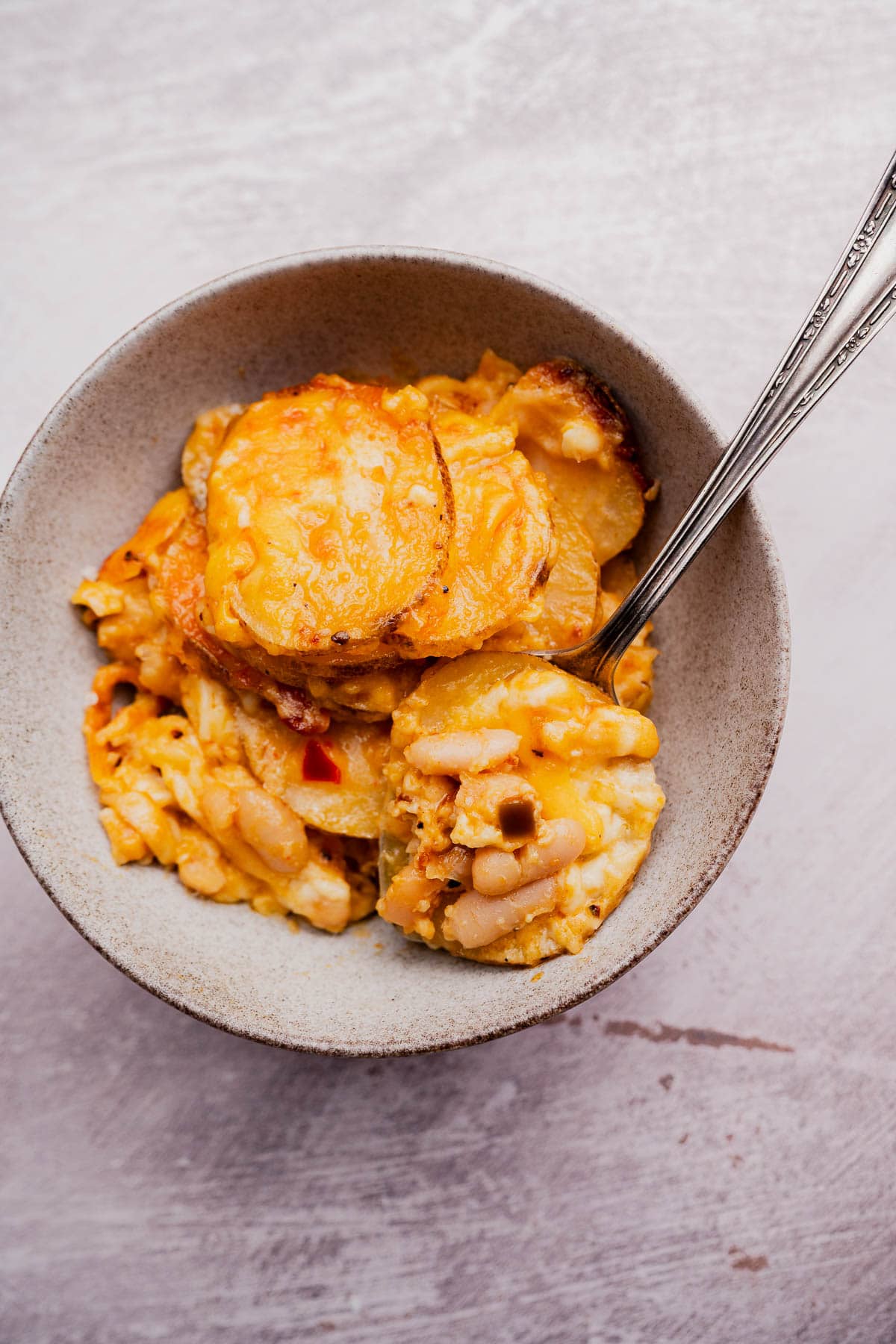 A bowl of buffalo potato casserole featuring sliced potatoes, white beans, and melted cheese, with a spoon resting inside.