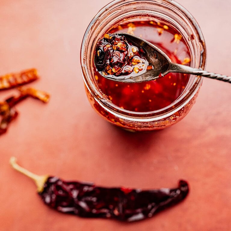 A jar of chili oil with a spoonful of fiery chili flakes held above it; dried red chili peppers rest on the vibrant red surface nearby.