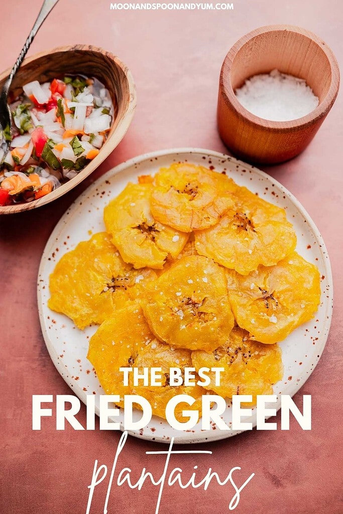 A plate of crispy patacones—fried green plantain slices—sprinkled with salt, next to a bowl of salsa and a small wooden bowl of salt, on a pink surface. Text reads: "The Best Fried Green Plantains.