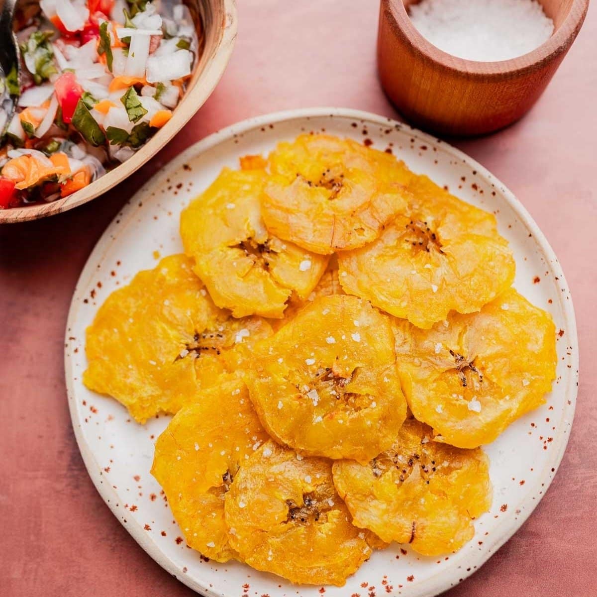 A plate of crispy patacones sprinkled with salt, served alongside a bowl of chopped vegetables and a small wooden container of salt nearby.
