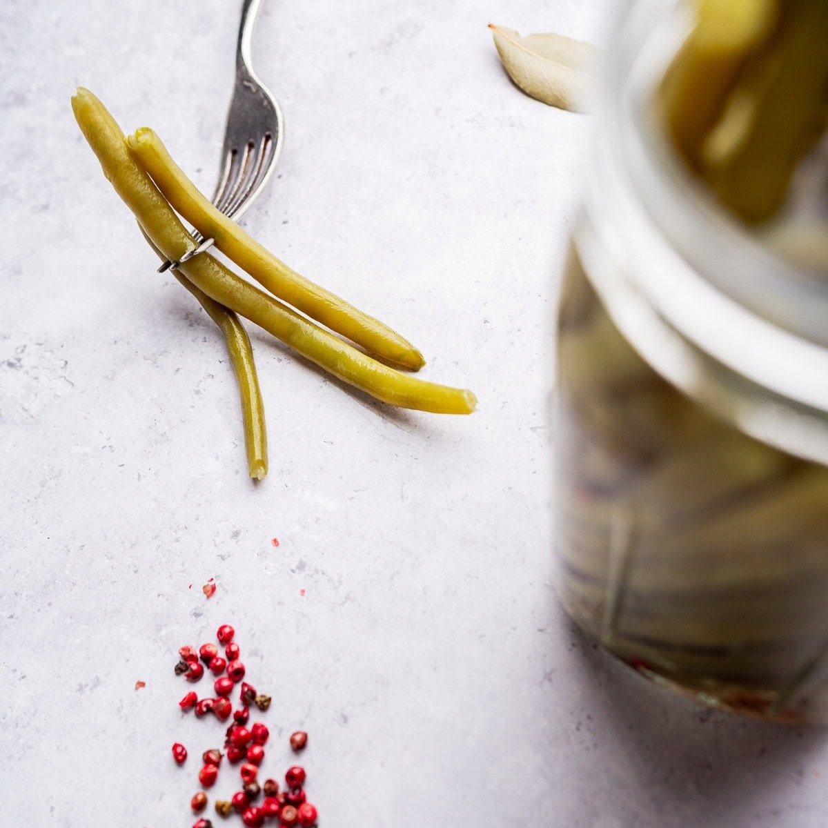 Three pickled green beans lie on a light surface, with a fork resting on them, a glass jar of pickled green beans, and scattered pink peppercorns nearby.