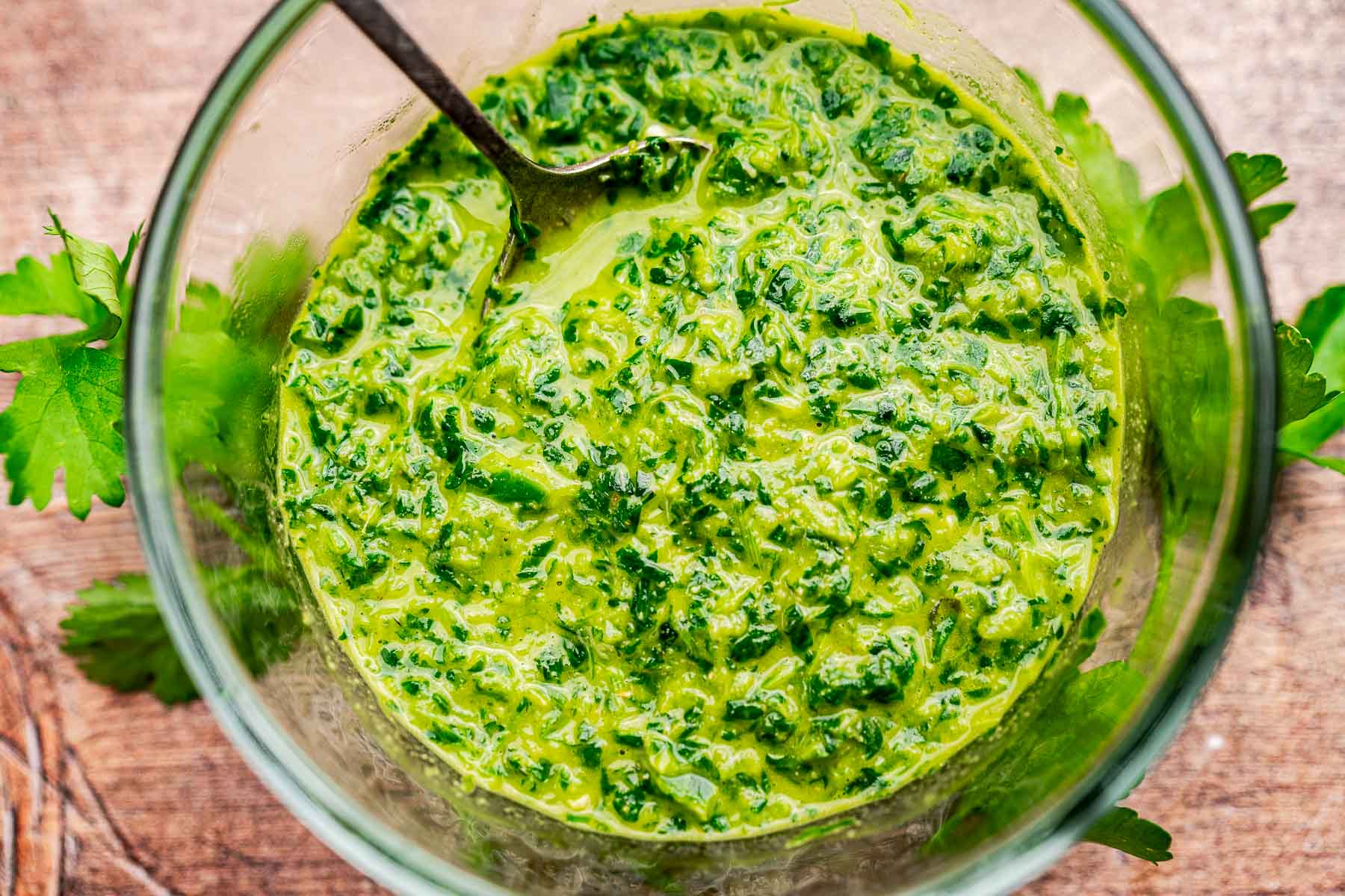 A glass bowl filled with green chimichurri sauce, resembling vibrant schug, with a metal spoon partially submerged in the sauce. Parsley leaves are visible next to the bowl on a wooden surface.