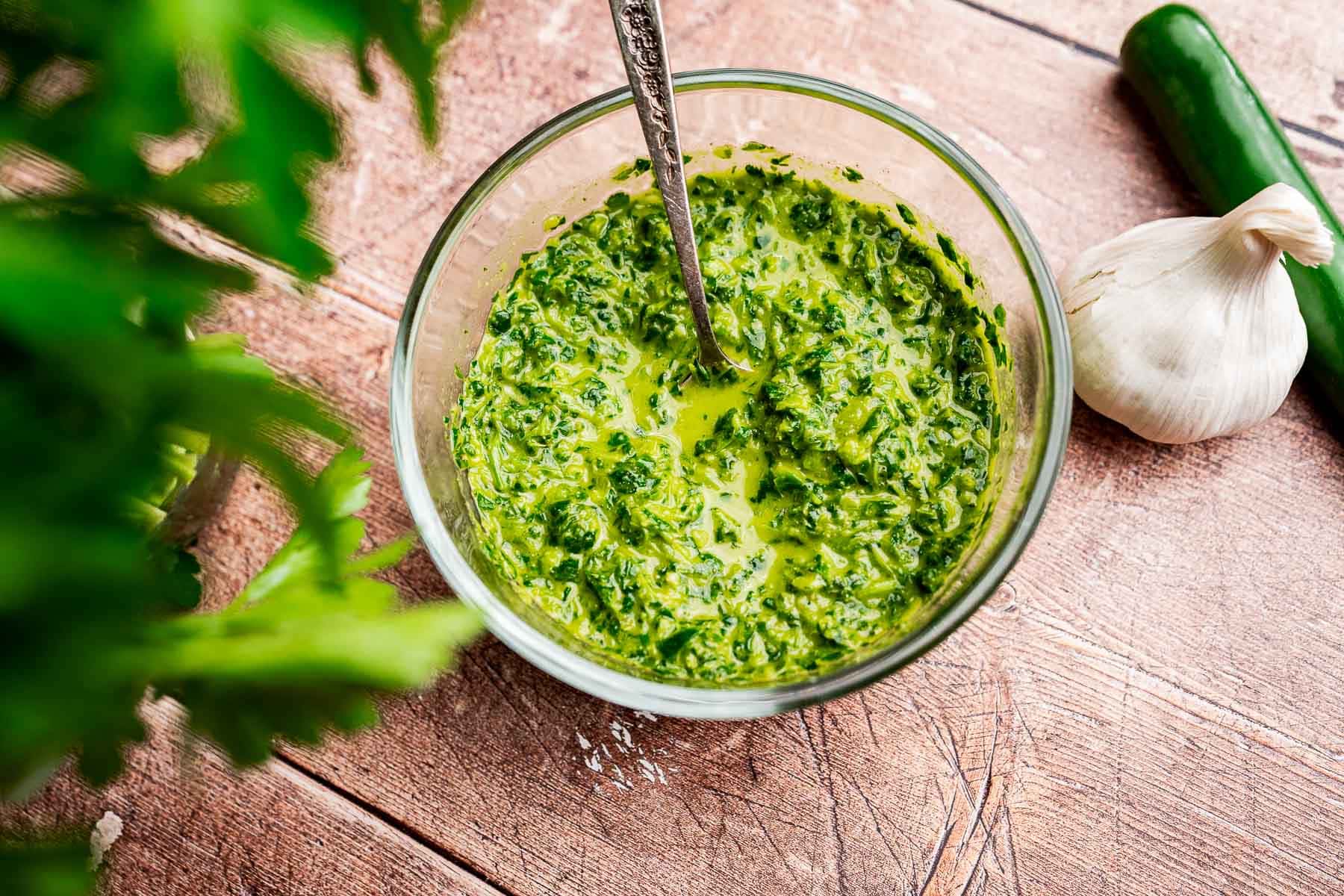 A bowl of vibrant green schug sauce with a spoon, placed on a wooden surface near a garlic bulb and a green chili pepper.