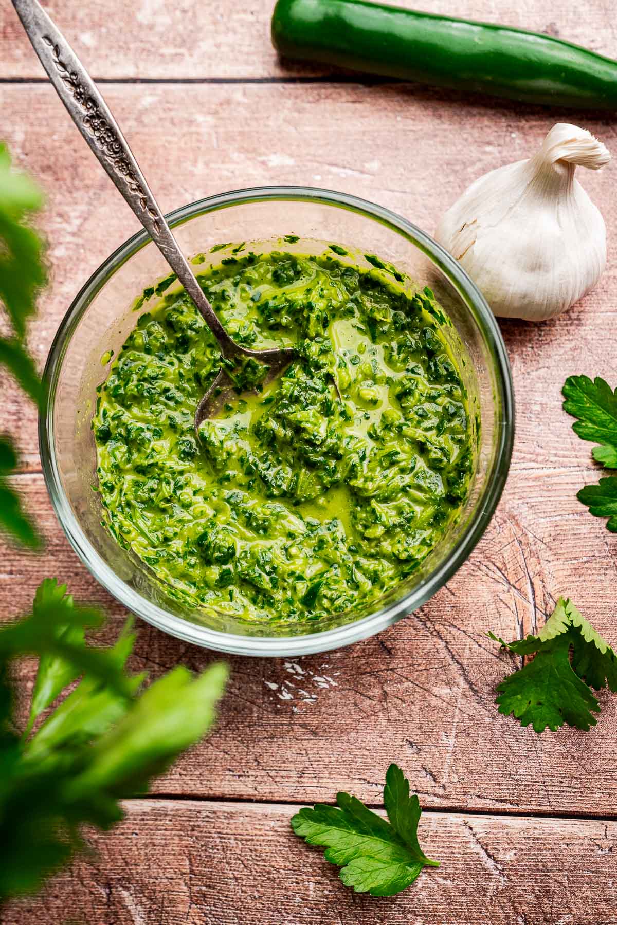 A glass bowl of vibrant green chimichurri sauce, reminiscent of schug, with a spoon, surrounded by a garlic bulb, green chili pepper, and parsley leaves on a wooden surface.