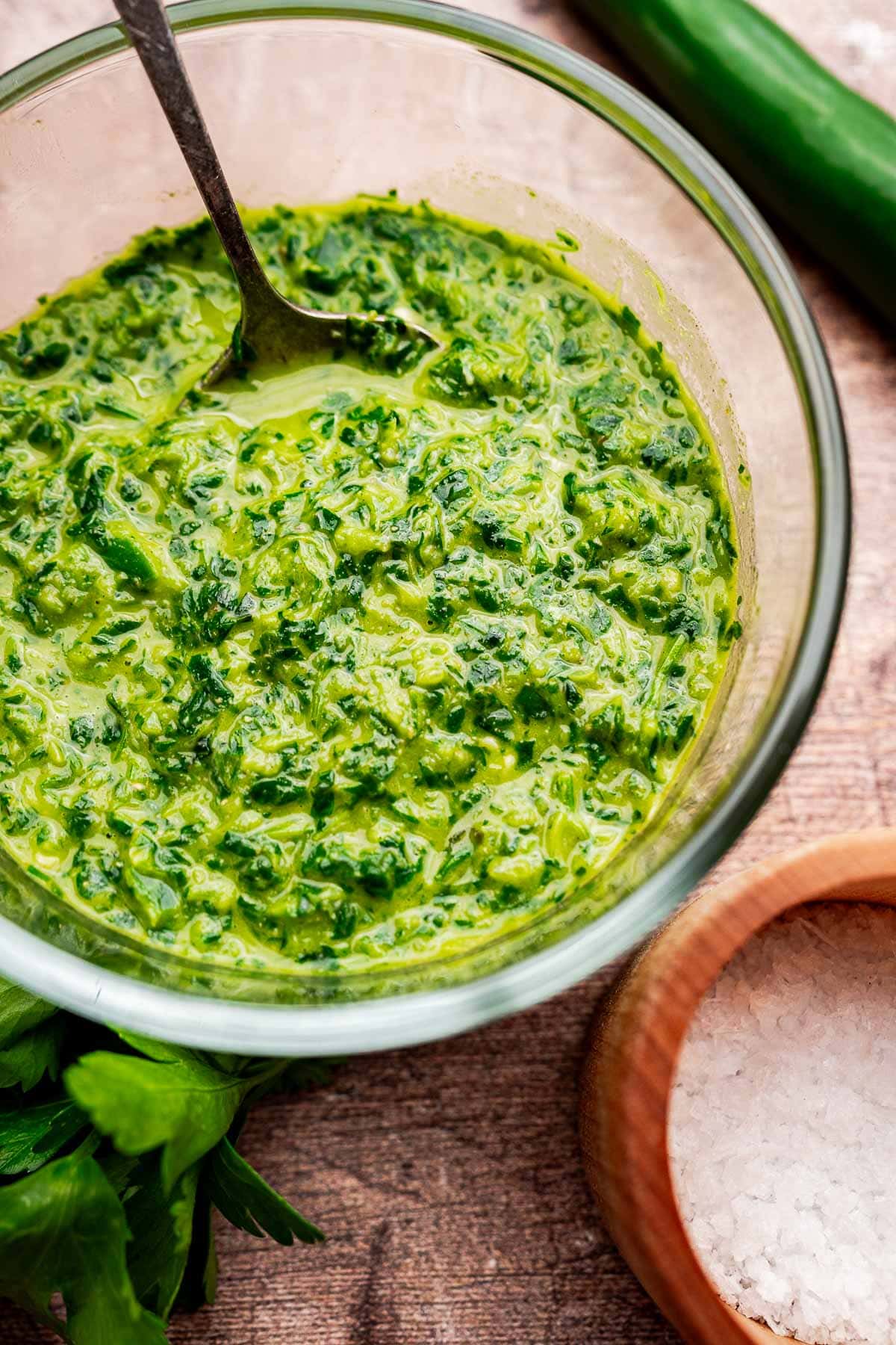 A glass bowl of green chimichurri sauce with a spoon, reminiscent of schug, sits next to a bowl of coarse salt and fresh herbs on a wooden surface.