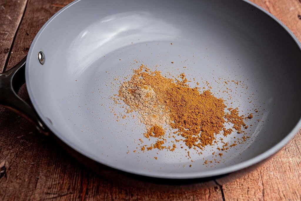 A gray frying pan on a wooden surface holds two small piles of ground spices, one light brown and one darker brown, perfect for adding bold flavor to schug or your favorite dishes.