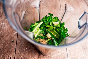 Fresh herbs, green chili, garlic cloves, and spices come together in a blender to create vibrant schug, ready to be mixed on a wooden surface.