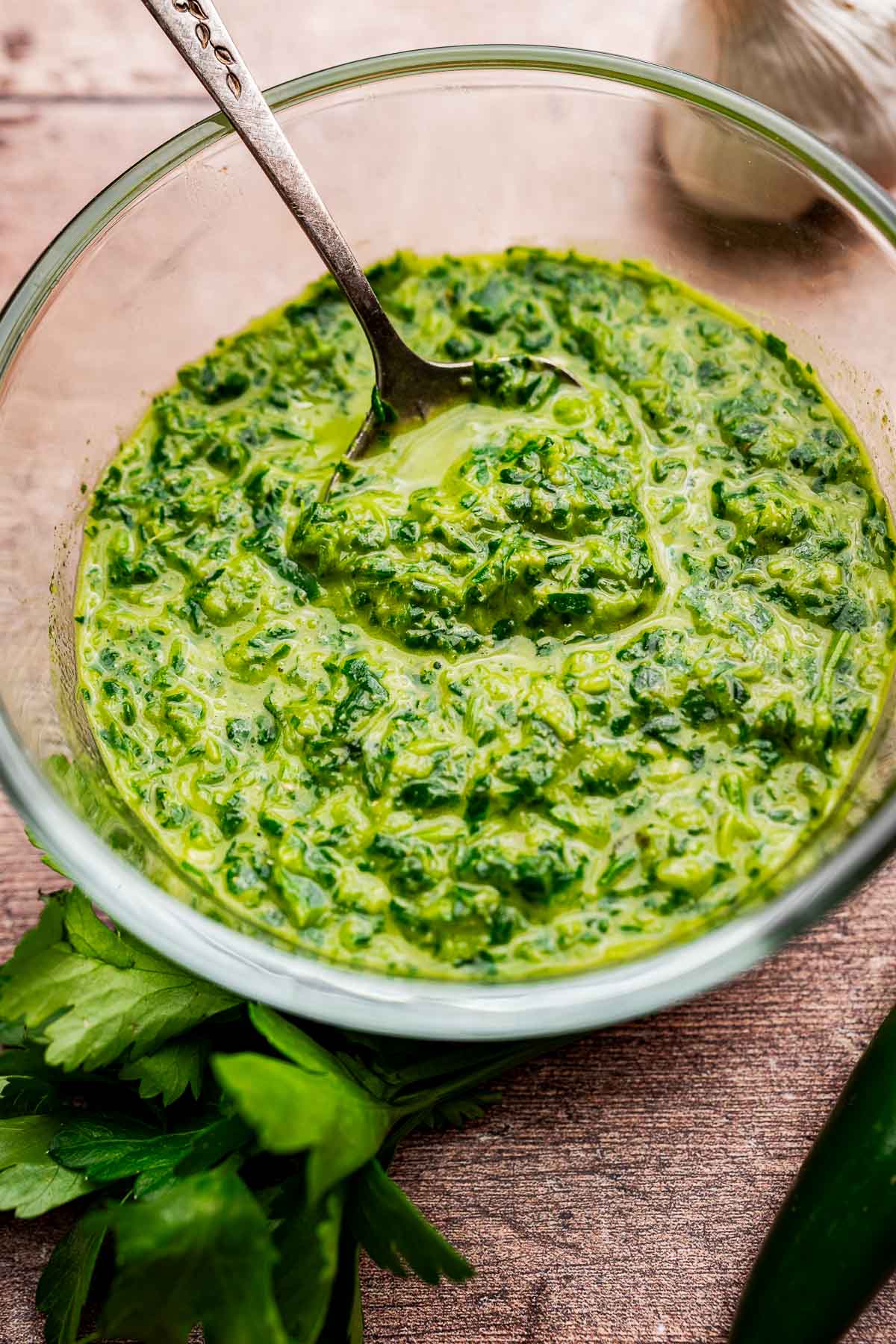 A glass bowl filled with vibrant green chimichurri and schug sauce, with a spoon inside and fresh herbs placed beside the bowl on a wooden surface.