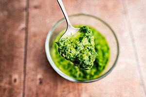 A spoonful of schug is held above a glass bowl filled with the vibrant green sauce on a wooden surface.