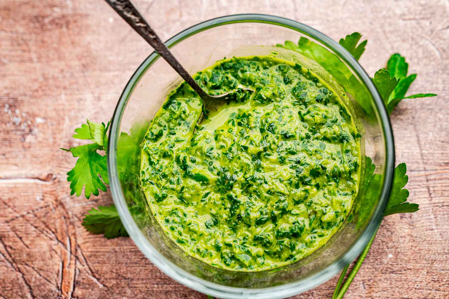 A glass bowl filled with vibrant green chimichurri sauce, blended with schug for extra heat, sits on a wooden surface. A spoon rests inside, and the sauce is garnished with fresh parsley.