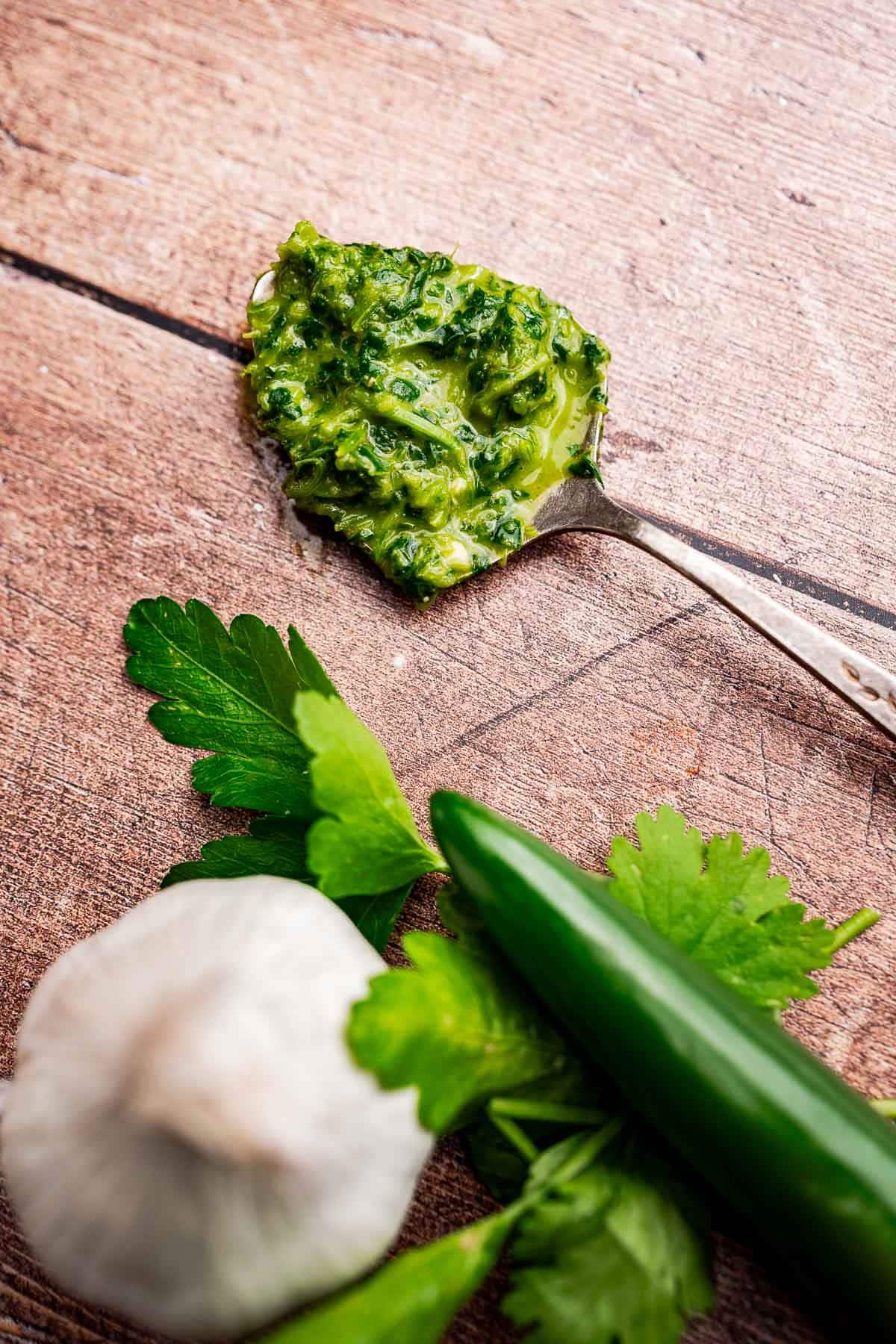 A spoonful of schug green sauce on a wooden surface beside a garlic bulb, a green chili pepper, and fresh parsley.