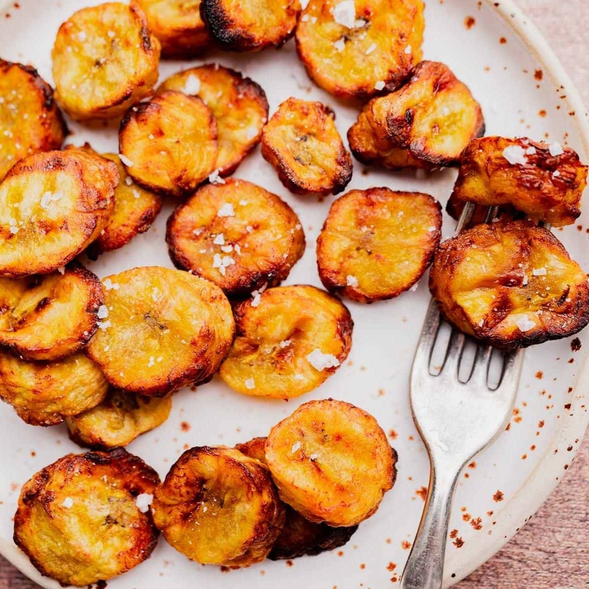 A white plate with slices of air fryer sweet plantains sprinkled with salt, next to a metal fork.
