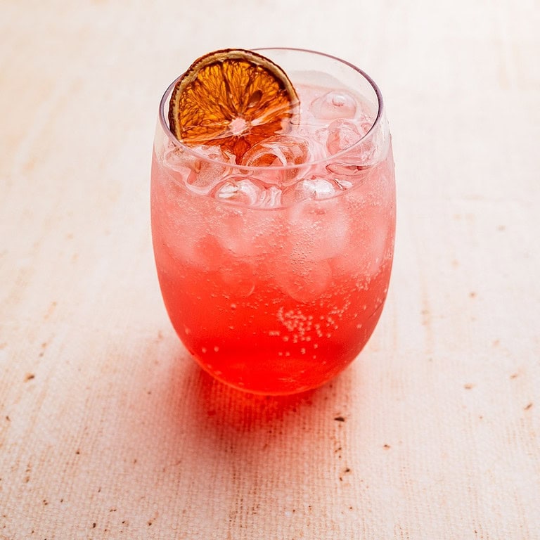 A glass of pink fizzy Aperol Spritz mocktail with ice cubes, garnished with a dried orange slice, on a light-colored surface.