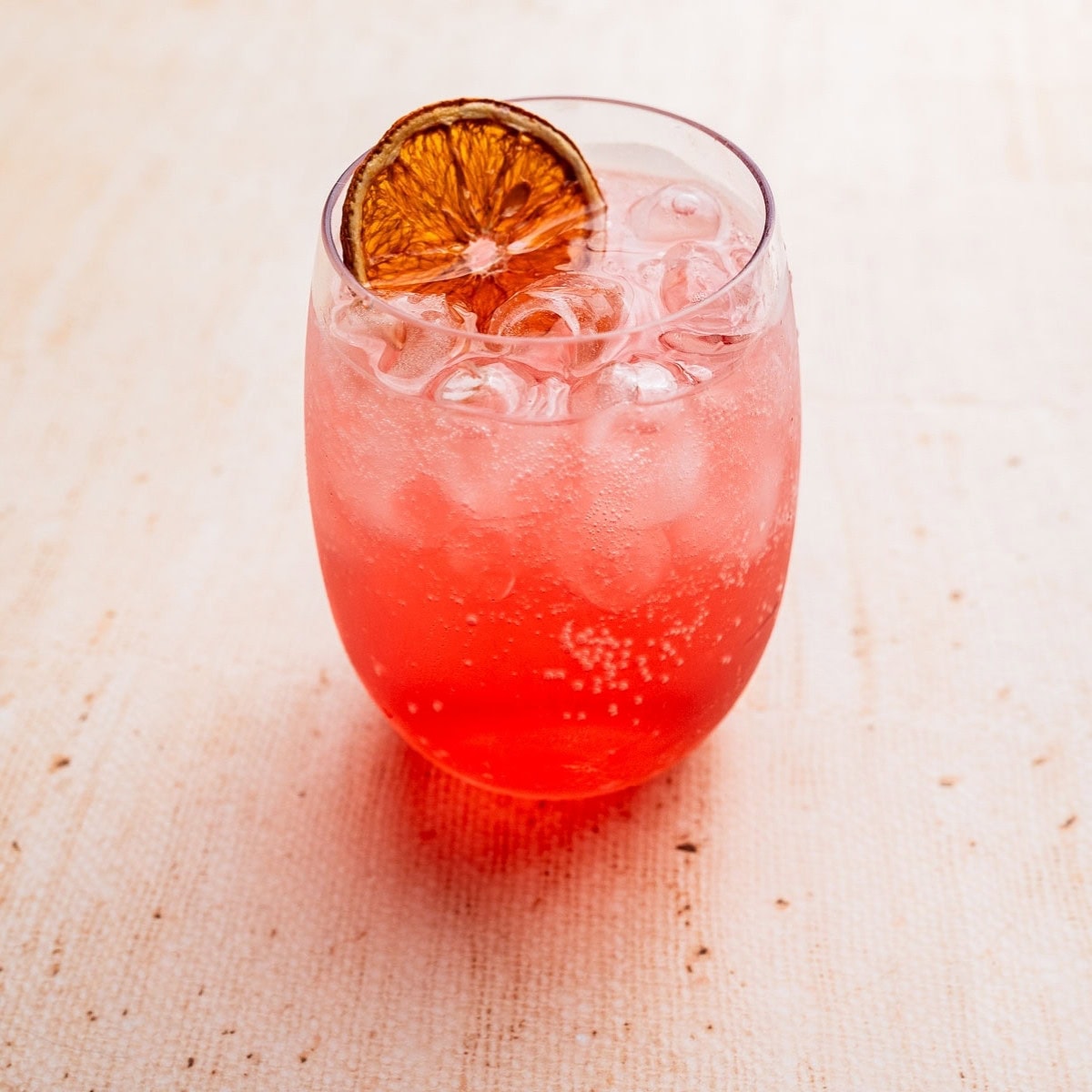 A glass of pink fizzy Aperol Spritz mocktail with ice cubes, garnished with a dried orange slice, on a light-colored surface.