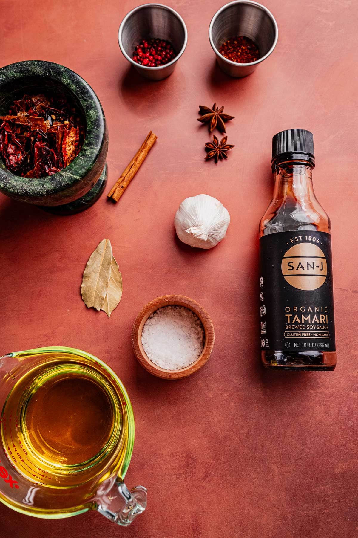 Ingredients for chili oil arranged on a countertop, including tamari sauce, oil, salt, spices, dried chilies in a mortar, and measuring cups with peppercorns and chili flakes.