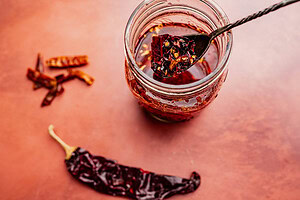 A jar of chili oil with a spoon inside, surrounded by dried chili peppers on a reddish surface, captures the bold essence of homemade chili oil.