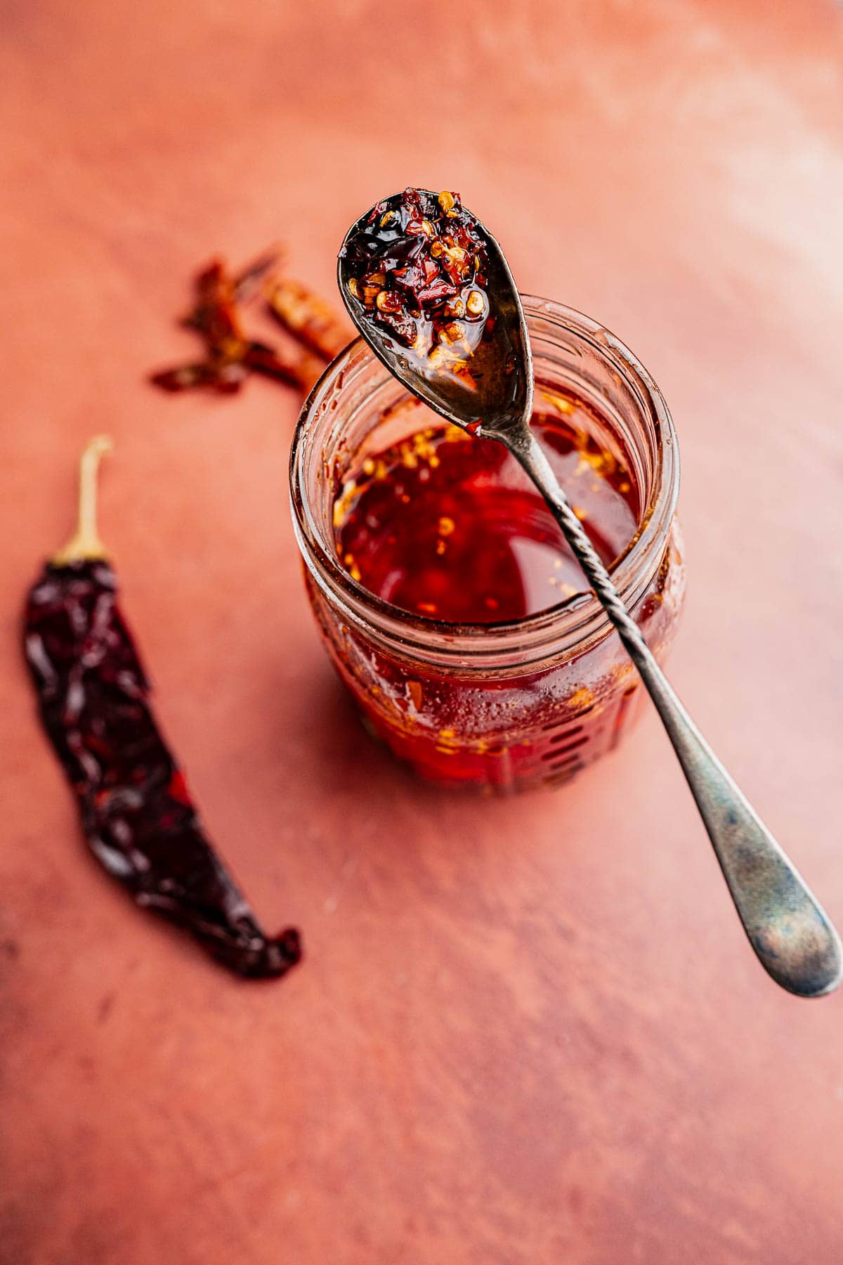 A spoon with chili oil and flakes rests on a jar of rich, aromatic chili oil, with dried chili peppers scattered on a reddish-brown surface nearby.