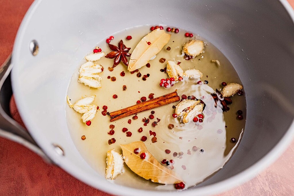 A saucepan containing water, bay leaves, star anise, cinnamon stick, sliced ginger, cardamom pods, pink peppercorns, and a hint of chili oil for a subtle kick.