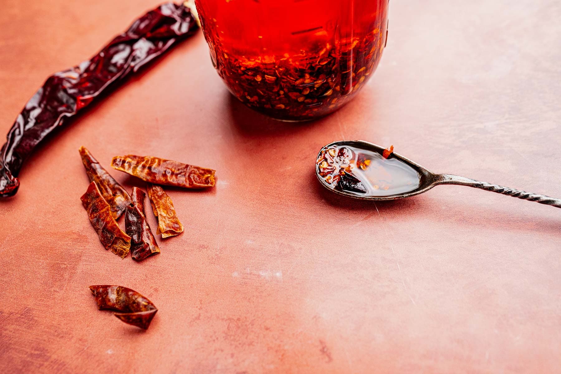 A spoon with chili oil, dried chili peppers, and a jar of homemade chili oil rest on a reddish surface.
