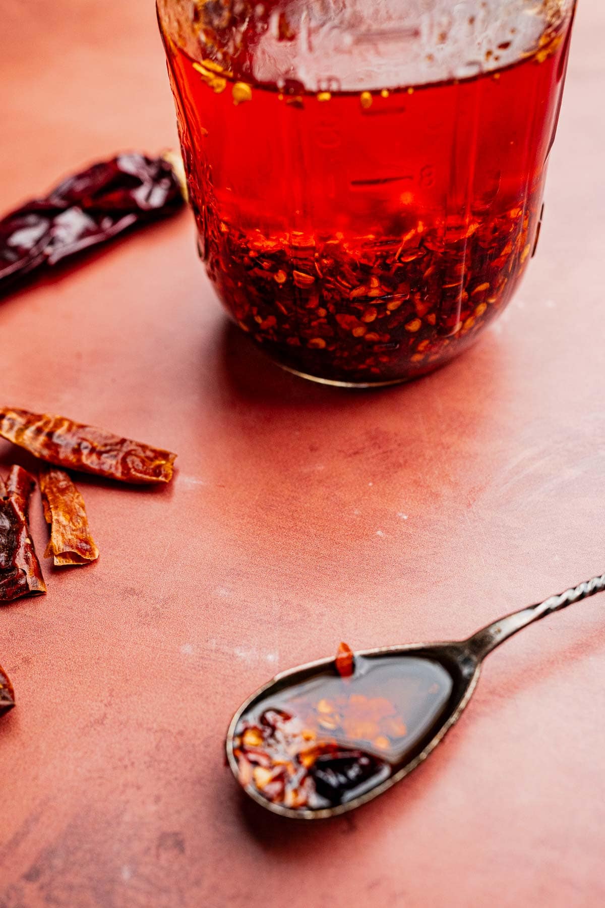 A jar of chili oil sits on a reddish surface beside dried chili peppers, with a spoon holding some vibrant chili oil and fiery flakes.