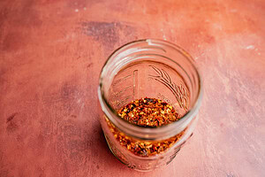 A glass jar containing crushed red pepper flakes sits on a reddish-brown surface, perfect for adding heat to dishes or infusing into chili oil.