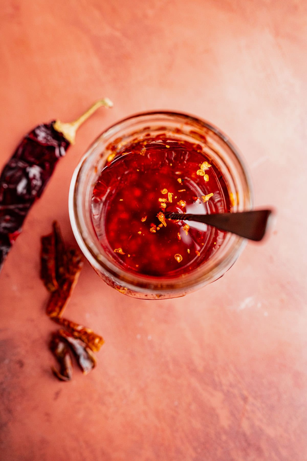 A jar of chili oil with a spoon inside sits on a reddish surface, surrounded by dried chili peppers. The vibrant chili oil glistens, inviting you to add a spicy kick to any dish.