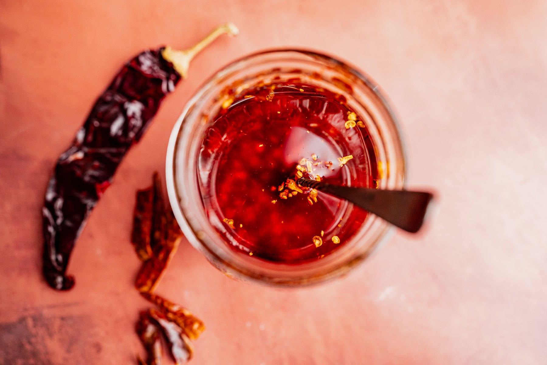 A glass jar of chili oil with a spoon, set on a reddish surface beside dried whole chilies.