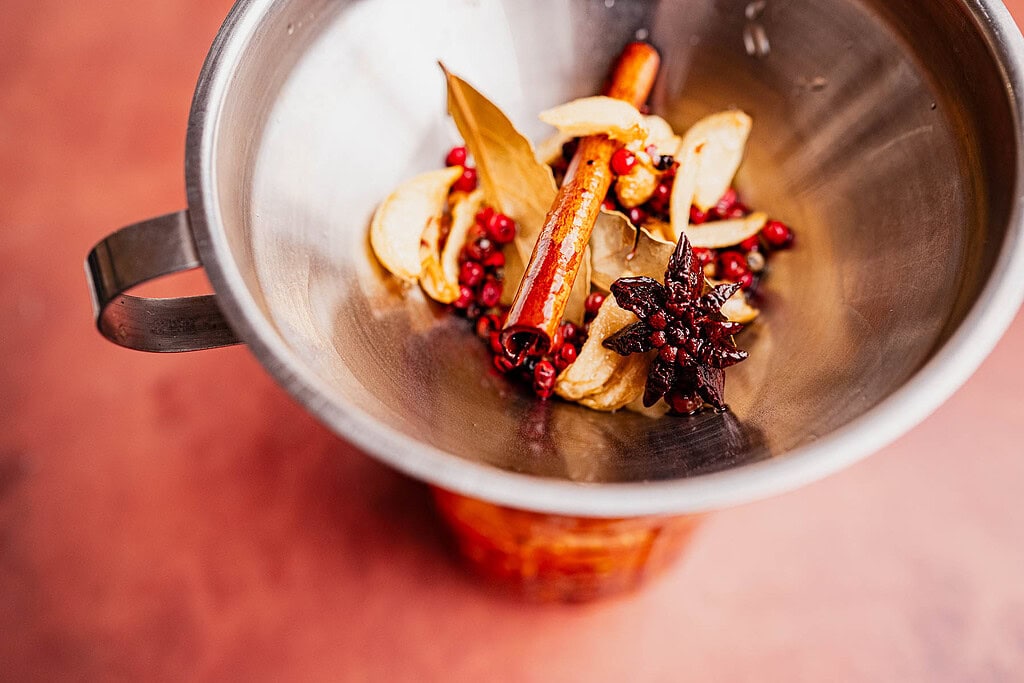 A metal bowl containing star anise, cinnamon sticks, bay leaves, peppercorns, dried ginger slices, and red spices for chili oil, viewed from above against a reddish surface.