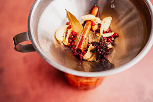 A metal bowl containing star anise, cinnamon sticks, bay leaves, peppercorns, dried ginger slices, and red spices for chili oil, viewed from above against a reddish surface.