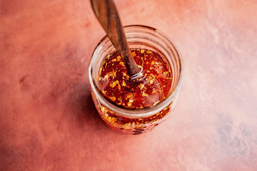 A glass jar of chili oil with red pepper flakes, containing a spoon, viewed from above on a reddish surface.