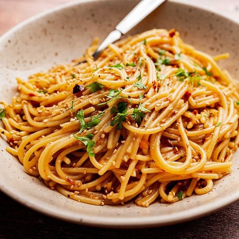 A bowl of chili oil pasta tossed with sauce, garnished with chopped parsley and sesame seeds, with a fork resting on the side.