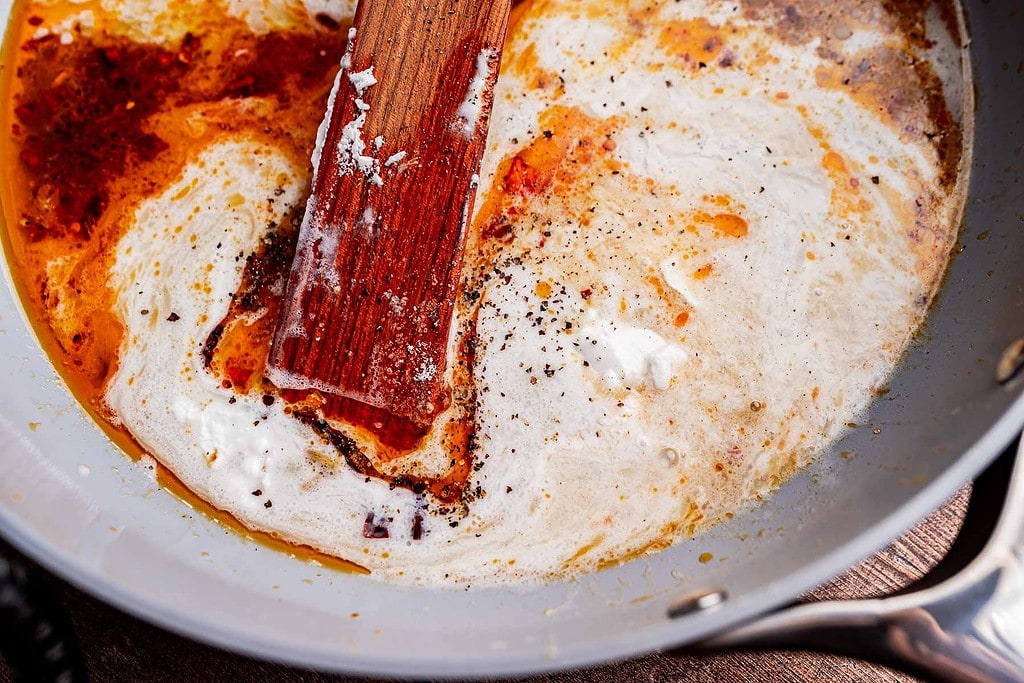 A wooden spatula stirs cream, spices, chili oil pasta, and seasoning in a skillet, creating a swirled mixture.