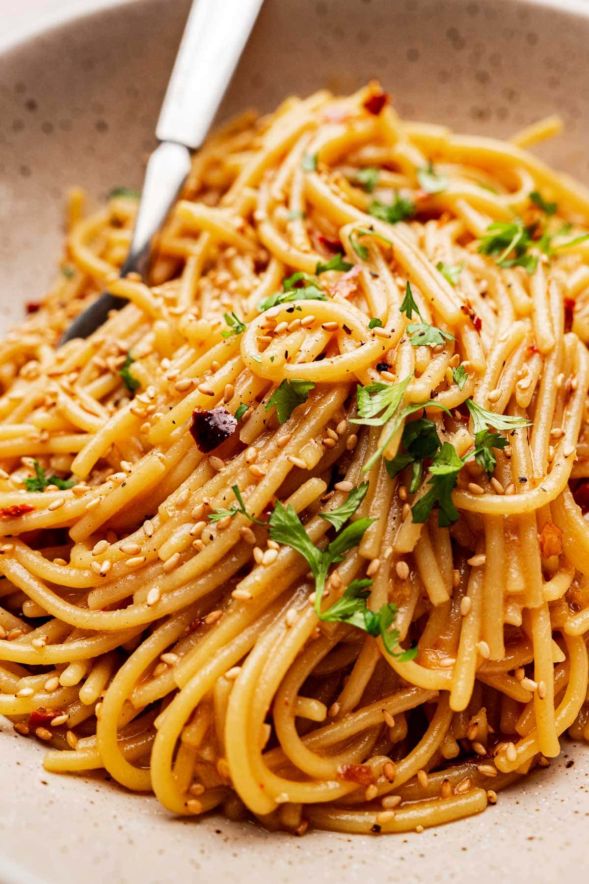 A bowl of chili oil pasta topped with chopped parsley, sesame seeds, and red pepper flakes, with a fork resting on the side.