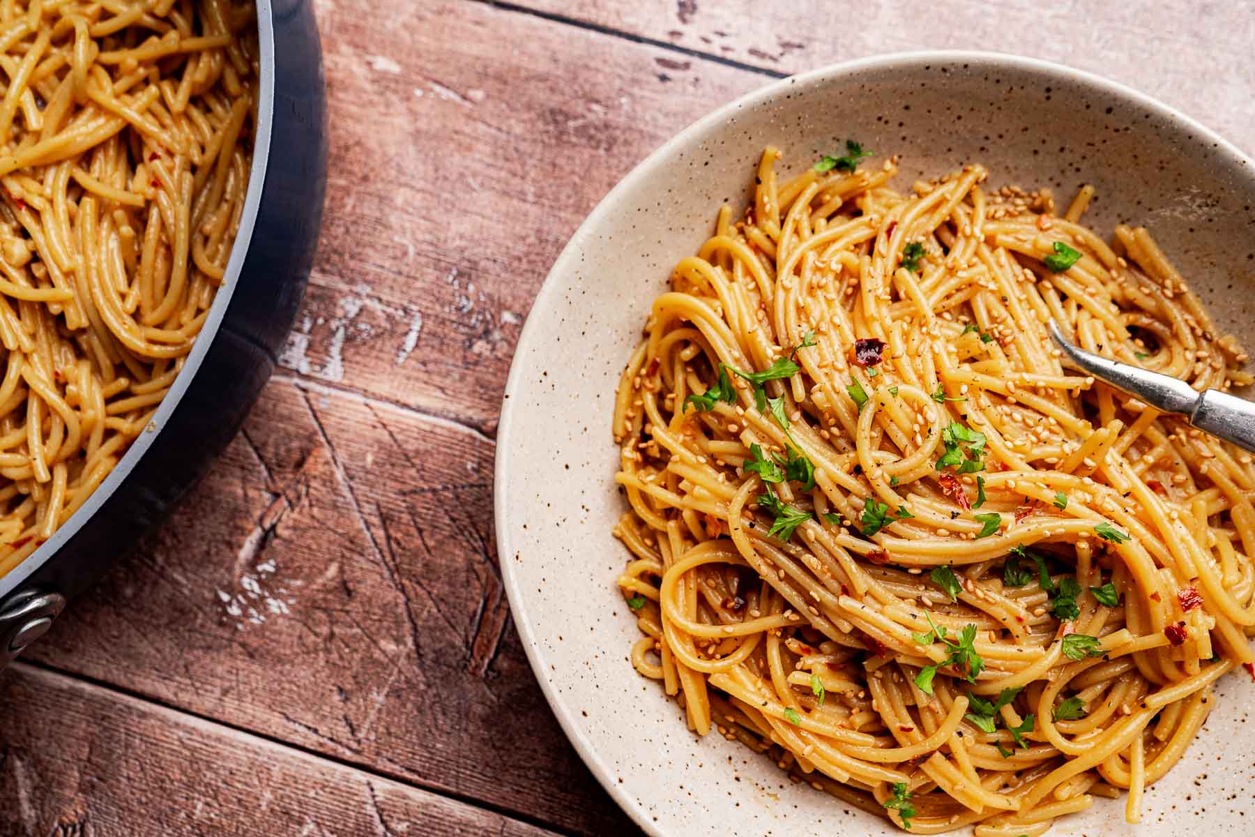 A plate of cooked spaghetti with herbs and chili oil pasta sauce sits on a wooden table next to a pan with more pasta.