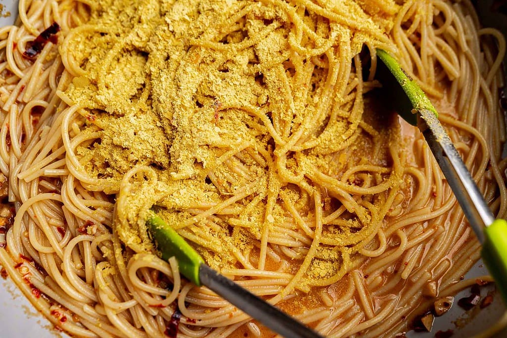 Close-up of cooked spaghetti being mixed with a yellow powdered seasoning and chili oil pasta using green-tipped tongs.