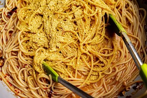Close-up of cooked spaghetti being mixed with a yellow powdered seasoning and chili oil pasta using green-tipped tongs.